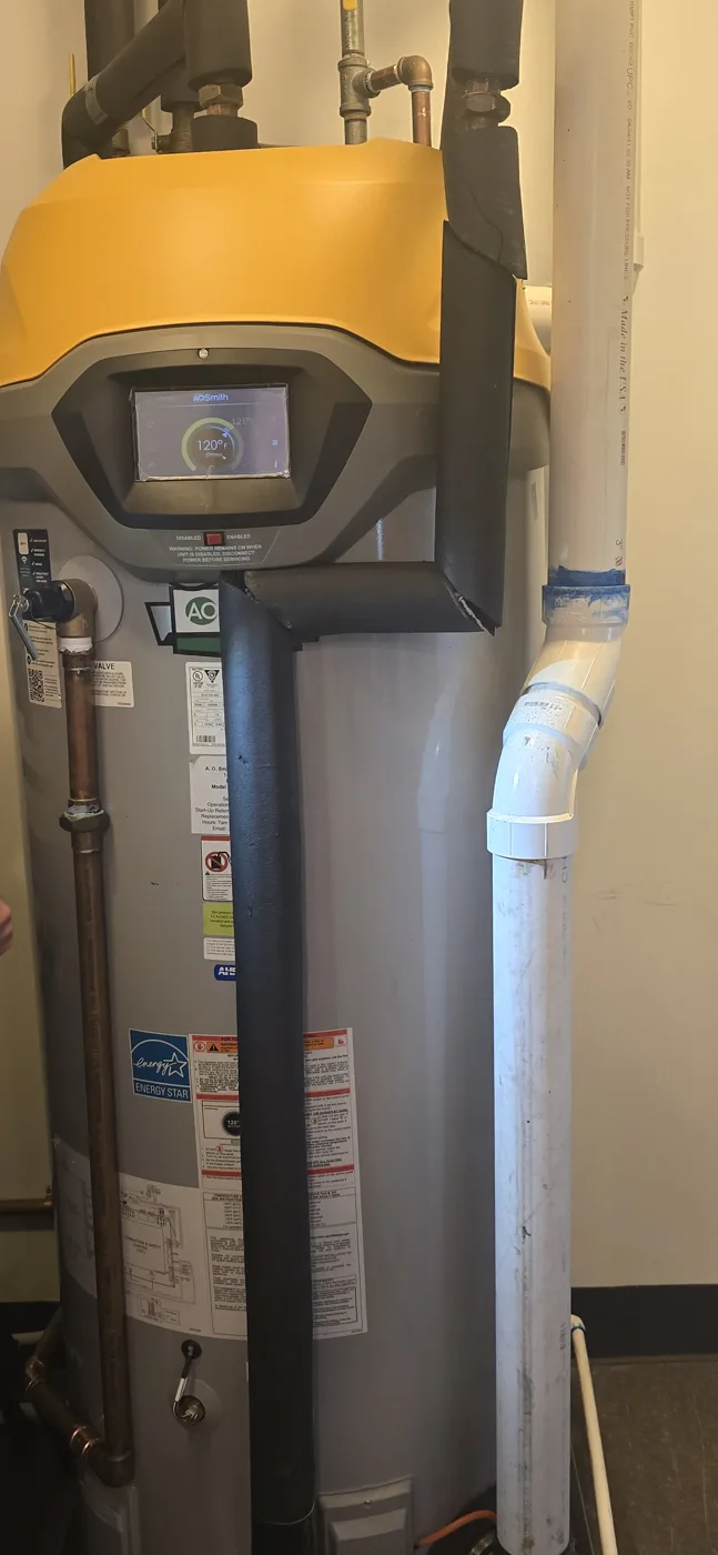 Heat-pump water heater with a gray tank, a yellow top and a lit touchscreen display, black insulated lines and a white PVC pipe beside it