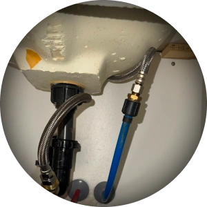 Underside of a sink basin with a black drain tailpiece, a braided steel supply hose and a blue supply line on a compression fitting